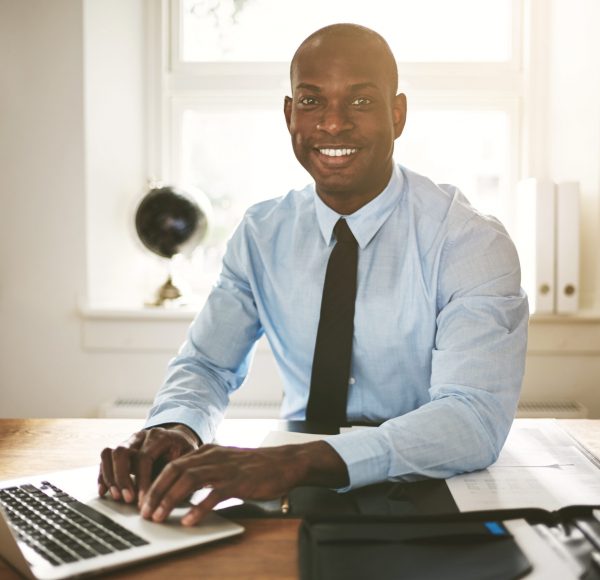 Successful young African businessman sitting at his desk in an office smiling and working on a laptop